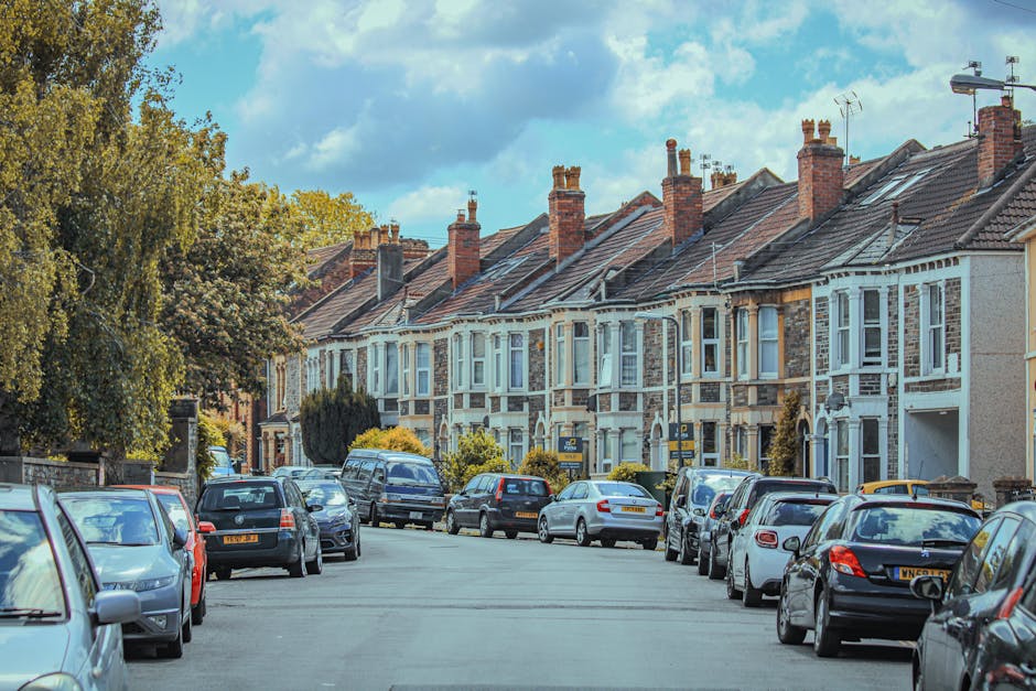A residential street lined with traditional Victorian terraced houses, featuring brick and stucco facades, bay windows, and pitched tiled roofs with multiple brick chimneys. The houses are closely pac